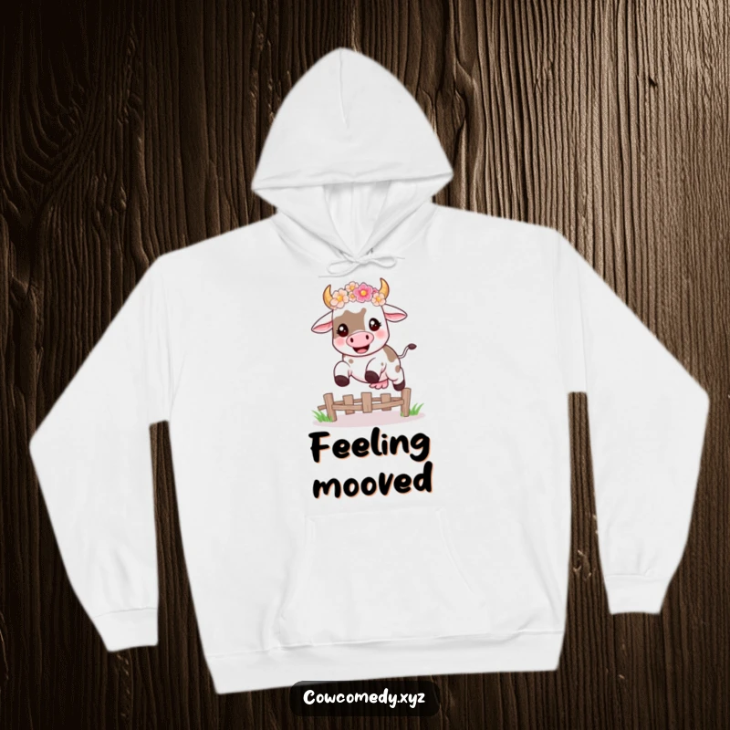 Funny hoodie featuring a happy kawaii cow wearing a flower crown, mid-leap over a fence, perfect for cozy, humorous wear.